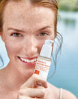 Woman holding skincare bottle in front of her cheek.